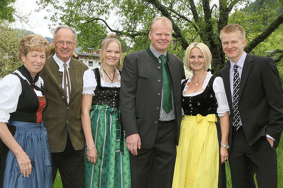 Sechs Personen in traditioneller bayerischer Tracht lächeln in der Natur. Im Hintergrund Bäume und ein Haus. Männer in Anzügen, Frauen in farbigen Dirndln.