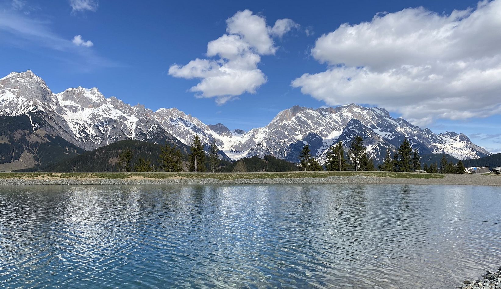 Ein klarer Bergsee mit schneebedeckten Bergen im Hintergrund und blauem Himmel. Am Ufer sind Kieselsteine und wenige Bäume zu sehen.
