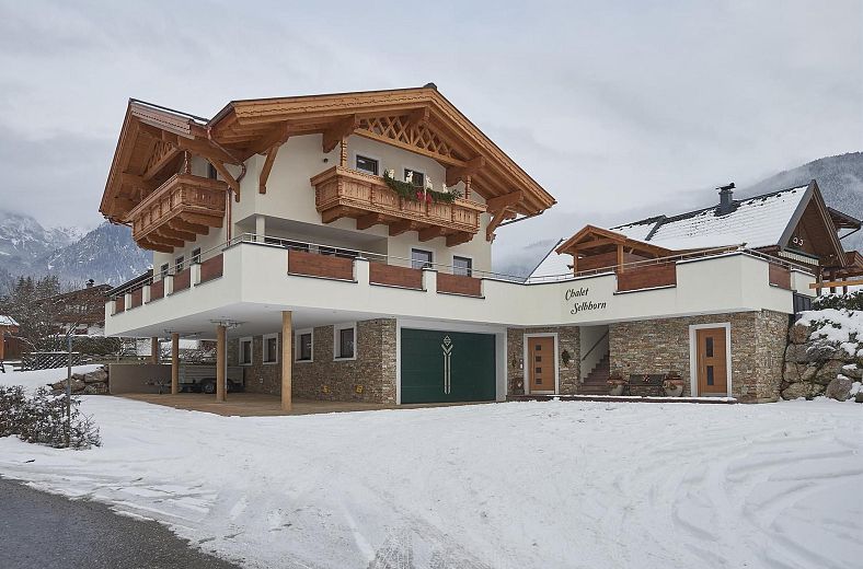 Ein zweigeschossiges alpenländisches Haus mit Holzelementen und Steinmauern, umgeben von Schnee, vor einer Bergkulisse.
