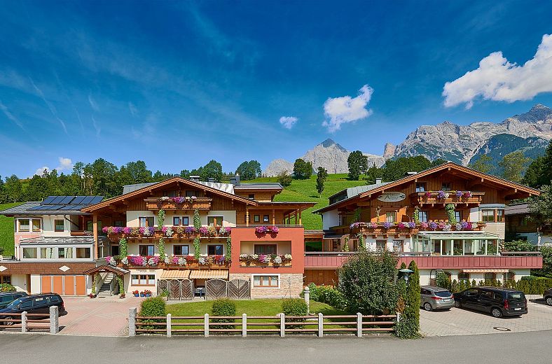 Ein traditionelles Alpenhotel mit bunten Fassaden und Blumenkästen. Im Hintergrund sind grüne Hügel und majestätische Berge unter einem klaren blauen Himmel zu sehen.