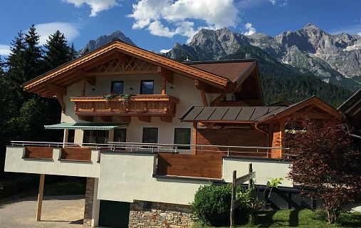 Ein charmantes Chalet in den Alpen mit Holzbalkonen vor dramatischen Bergen und blauem Himmel mit Wolken. Die Umgebung ist bewaldet und grün.