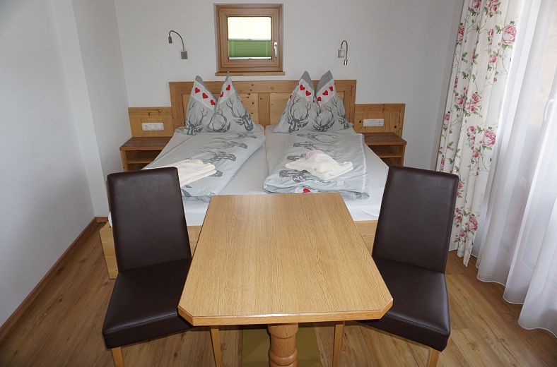 Ein einfach eingerichtetes Doppelzimmer mit zwei Betten, einem Holztisch und zwei Stühlen. Die Wände sind weiß, und eine kleine Fensteröffnung ist sichtbar.