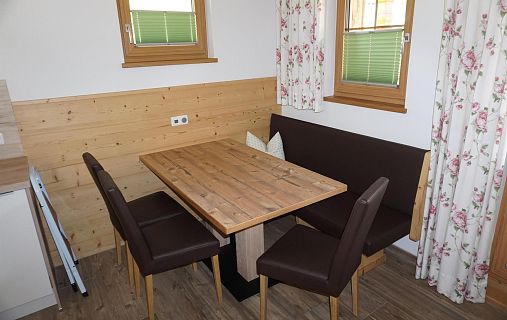 Essbereich mit Holztisch, gepolsterten Stühlen und L-förmiger Bank. Wände mit Holzverkleidung, Fenster mit geblümten Vorhängen, gemütliches Ambiente.