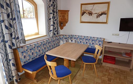 Kleine Essecke mit Holztisch und blauen Polsterstühlen. An der Wand ein Bild mit Händen, die sich berühren, inspiriert von Michelangelo. Helles Fenster.