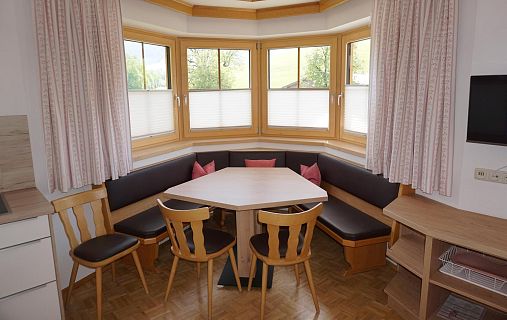 Eine gemütliche Essecke mit großen Fenstern, einem Holztisch und Polsterbänken. Die hellen Vorhänge und warmen Holztöne schaffen eine einladende Atmosphäre.