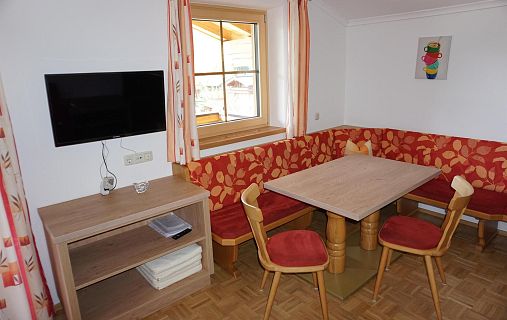 Ein Essbereich mit Eckbank und Holztisch, roter Polsterung, Fenster mit orangegestreiften Vorhängen und kleinem Wandfernseher.