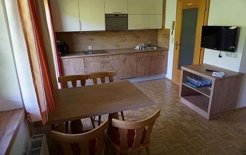 Kleine Küche mit Holzmöbeln, Tisch und Stühlen. Moderne Geräte eingebaut in eine Küchenzeile. Fenster mit Vorhängen und ein kleiner Fernseher an der Wand.