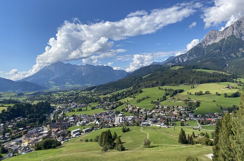 Blick auf eine grüne Alpenlandschaft mit einem Dorf, umgeben von hohen Bergen und blauem Himmel mit weißen Wolken.