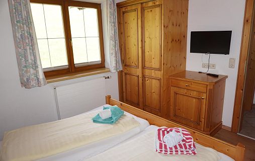 Schlafzimmer mit Holzschrank, Kommode, Bett mit Handtüchern und kleinem Fernseher. Fenster mit Gardinen im Hintergrund.