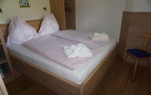 Ein Hotelzimmer mit einem großen Doppelbett, schlicht eingerichtet mit Holzmöbeln und rosa gestreifter Bettwäsche. Ein Stuhl steht neben dem Bett.