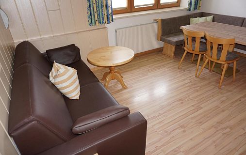 Ein Wohnzimmer mit braunem Ledersofa, rundem Holztisch, Esstisch mit Stühlen und Fenster. Holzboden und Gardinen ergänzen das warme Ambiente.