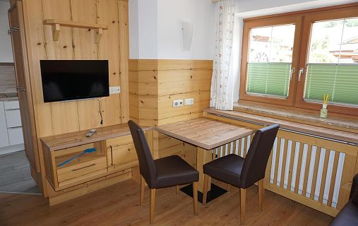 Eine Holzecke mit kleinem Tisch, zwei Stühlen und einem Fernseher an der Wand. Ein Fenster mit grünen Jalousien und ein Pflanzenblatt in der Ecke.