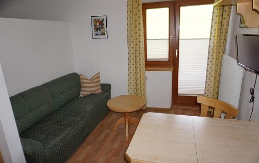 Kleines Wohnzimmer mit grünem Sofa, Holztisch, Fernseher an der Wand, und Gelbvorhängen an einem Fenster.