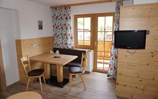Wohnzimmer im Alpenstil mit Holztisch, Stühlen, Fernseher und Blumenmuster-Vorhängen. Balkontür öffnet sich zu einem Holzbalkon.