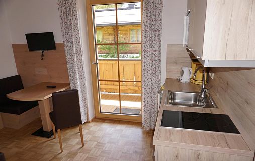 Kleine Küche in einer Ferienwohnung mit Herd, Spüle, Esstisch und Zugang zu einem Balkon durch eine Glastür. Helle Holzverkleidung dominiert das Interieur.