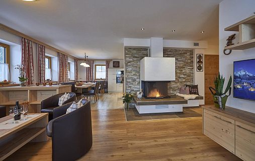 Ein Wohnzimmer mit Holzböden, modernem Kamin, gemütlichen Sitzmöbeln und Dekoration im alpinen Stil. Große Fenster sorgen für viel Tageslicht.